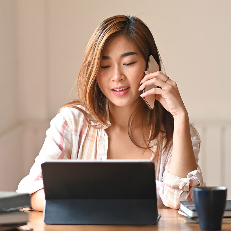 A woman sits at a table, working on a laptop while listening to on-hold messaging